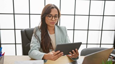 Ciddi ve başarılı genç İspanyol kadın ofis masasında dizüstü bilgisayarla ve dokunmatik bilgisayarla patronluk taslıyor..