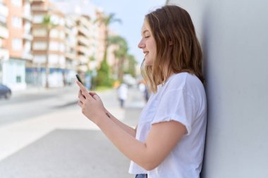 Beyaz bir kadın sokakta akıllı telefon kullanarak gülümsüyor.