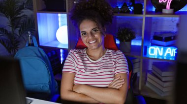 Smiling young hispanic woman with curly hair, arms crossed, in dark modern gaming room at night. clipart