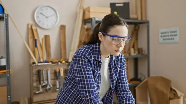在由工具和木料环绕的木工车间里戴安全眼镜的专注年轻女性 — 图库照片