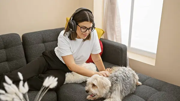Hispanic woman with headphones petting dog on a couch in a cozy living room