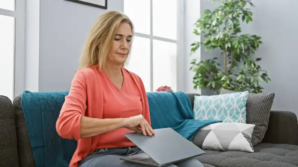 stock image Mature blonde woman closing laptop in a modern living room, depicting daily life and technology use.