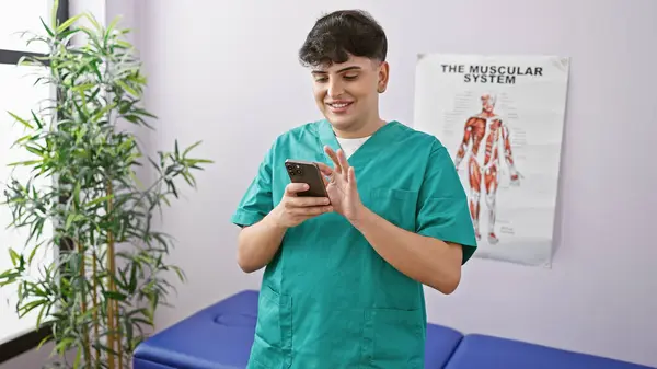 A young man in medical scrubs uses a smartphone in a clinic room with an anatomical poster on the wall.
