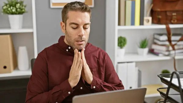 stock image A contemplative young hispanic man with a beard in an office setting appears focused or stressed.