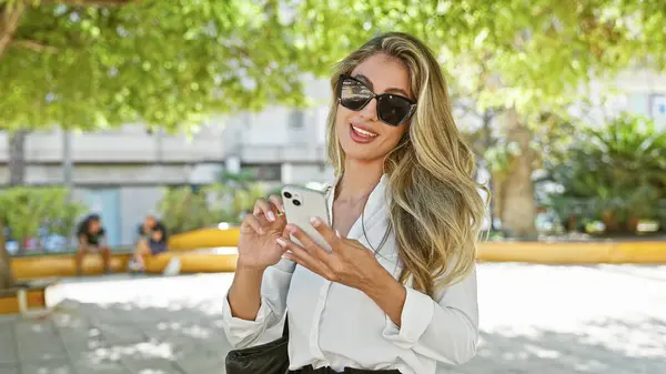 一位快乐 迷人的金发年轻女子在智能手机上愉快地输入手机短信 一边沉浸在网上对话中 一边泡在城市公园的绿地里 — 图库照片