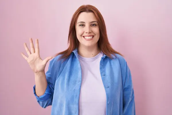 ピンクのバックグラウンドの上に立っている赤い髪の若いヒスパニックの女性は自信と幸せに微笑んでいる間指番号5を指で指を指さす — ストック写真