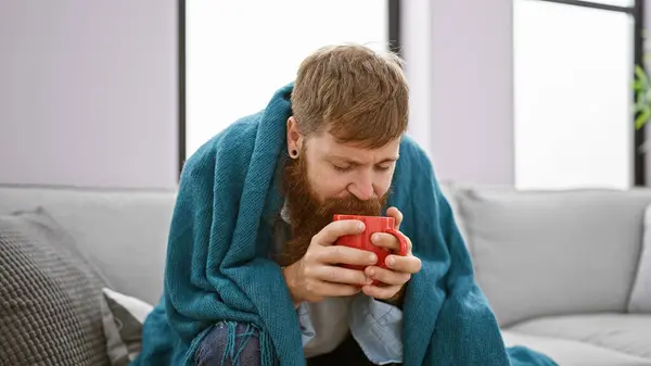 Chilled to the bone, young, handsome redhead man drinks hot coffee, seeking comfort and warmth under a cozy blanket on the sofa, battling the cold winter indoors at home
