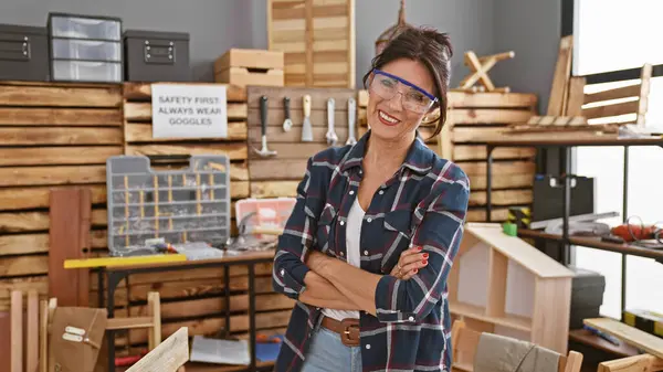 Smiling woman with safety goggles stands confidently in a cluttered carpentry workshop, embodying skilled labor.