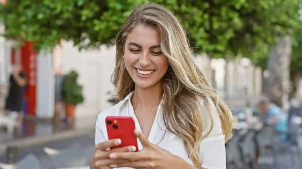 美しい若いブロンドの女性が幸せに触れ 彼女のスマートフォンでのエキサイティングなオンライン会話に深く魅了され 都市の通りで屋外で笑顔 陽気なバイブを放射します — ストック写真