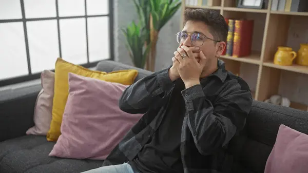 Stock image A young hispanic man with a moustache sitting indoors, covering his mouth with his hands, in a thoughtful pose on a sofa at home.