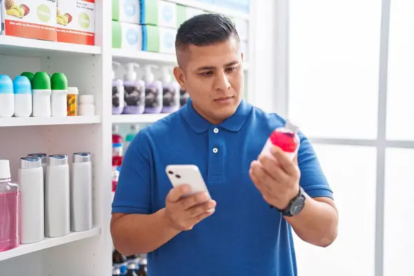 Young latin man customer using smartphone holding medicine bottle at pharmacy