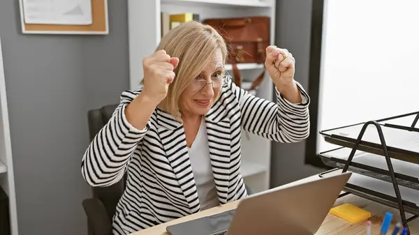 兴奋的金发女人带着手提电脑和笔记本在办公室的工作站庆祝成功 — 图库照片