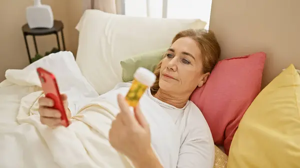 Зрелая Женщина Осматривает Бутылку Лекарствами Лежащую Спальне Смартфоном Подушками Тумбочкой — стоковое фото