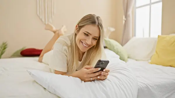 年轻而自信的金发女子在床上享受着早晨的时光 在舒适的卧室里用智能手机愉快地微笑着 — 图库照片