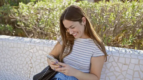 ベンチの日光公園でスマートフォンを使用してブルネット髪の若い女性を笑顔に — ストック写真