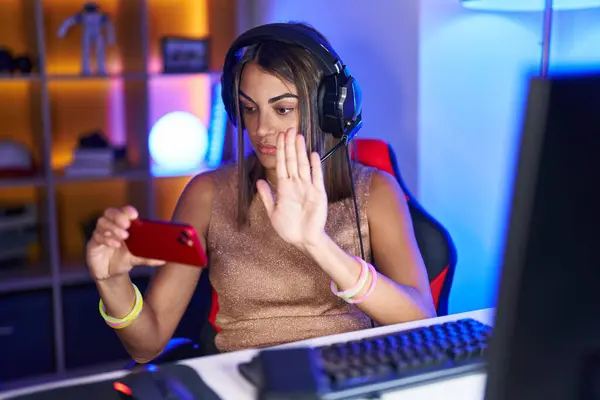 stock image Young hispanic woman playing video games with smartphone with open hand doing stop sign with serious and confident expression, defense gesture 