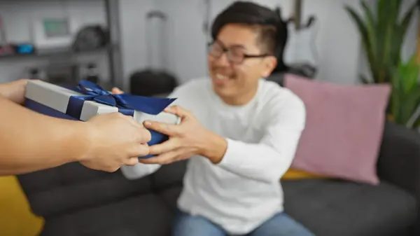 An asian man excitedly receiving a gift from another person in a cozy living room setting.