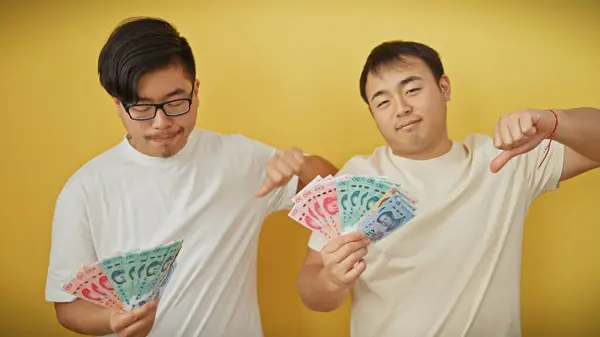 Two asian men displaying thumbs down gesture while holding chinese yuan against a yellow background.