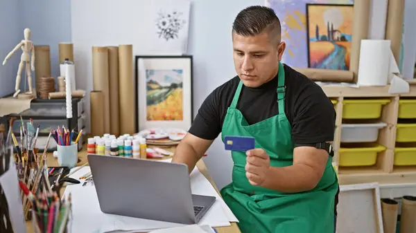 Hardworking latin artist, young man handling the responsibilities of his career with laptop and credit card in hand at art studio