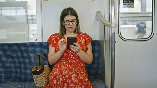 美しい若いヒスパニック系女性がメガネを寄贈し 彼女が都市生活を受け入れて 空の地下鉄で彼女のスマートフォンを使って一人で座って 彼女の携帯電話に魅了された肖像画 — ストック写真