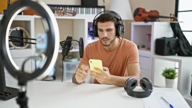 Müzik stüdyosunda mikrofon ve kulaklıkla akıllı telefon kullanan sakallı yakışıklı bir İspanyol.