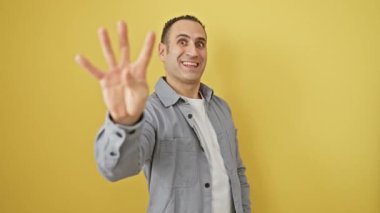 Tişörtlü neşeli genç İspanyol adam, mutlu sarı bir arka planda, kendinden emin ve izole bir şekilde, dört el işareti gösteriyor.