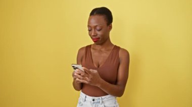 Pozitif, kendinden emin Afro-Amerikan kadını akıllı telefonuyla sevinçle gülümsüyor, izole edilmiş sarı arka planda neşeli mesajlar atıyor.