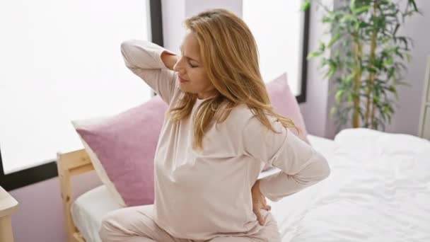 パジャマの痛みを伴う若い女性は 屋内ベッドに座って 不快感や腰痛を描写しながら背中を把握する — ストック動画