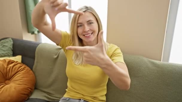 笑着的金发年轻女人在家里捕捉着自己的快乐 用手指做了一个相框 体现了摄影的创造力 — 图库视频影像