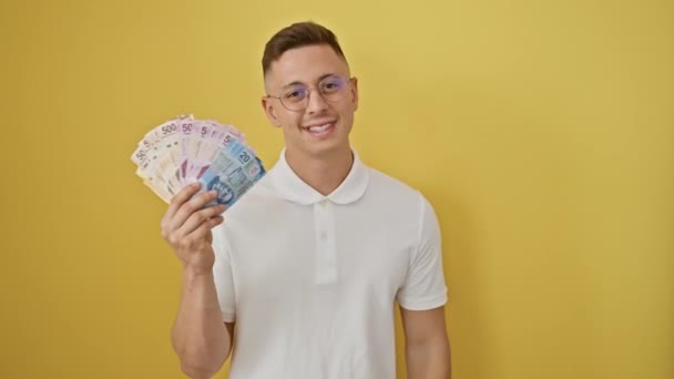陽気な若いヒスパニックの男性は喜んでメキシコのペソ紙幣の束に親指で指差し 孤立した黄色の背景に対して口を開いた笑顔で — ストック動画