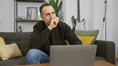 Düşünceli İspanyol adam rahat oturma odasında dizüstü bilgisayarla çalışırken gözlük tutuyor..