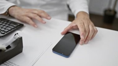 Ofiste çalışan profesyonel bir adam akıllı telefon, klavye ve belgelerle iletişim kurarak modern iş yeri teknolojisi kullanımını resmediyor..
