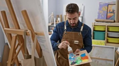 Rahatlamış genç İspanyol bir adam, yakışıklı sakallı bir ressam portresi, resim stüdyosunda fırçalarla resim yapmaya odaklanmış, tuval başyapıtını mükemmelleştiriyor.