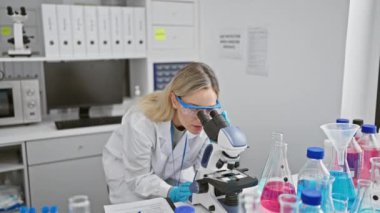 Modern bir laboratuvar ortamında mikroskop kullanan odaklanmış sarışın bir bilim kadını..