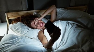Yatakta uzanmış gülümseyen genç bir kadın, gece evde akıllı telefon kullanıyor.