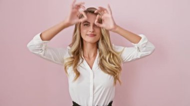 Genç sarışın kadın ayakta dikiliyor, dürbün gibi jest yapıyor, gözleri parmaklarından bakıyor. Çılgın bir ifade. izole edilmiş pembe arkaplan