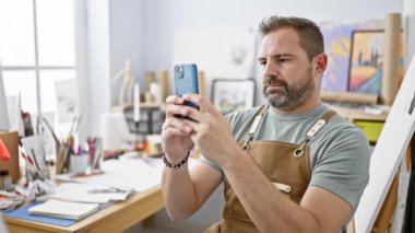 Parlak bir sanat stüdyosunda akıllı telefon kullanan önlüklü, orta yaşlı yakışıklı bir adam..