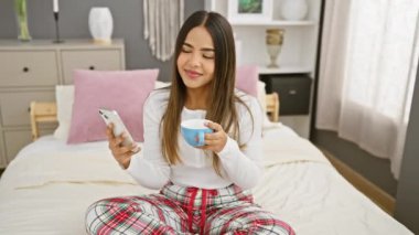 Pijamalı genç bir İspanyol kadın modern bir yatak odasında akıllı telefonunu kontrol ederken sabah kahvesinin tadını çıkarıyor..