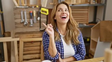 Neşeli genç Latin kadın, marangozluk atölyesinin yan tarafını göstererek, yüzünde güzel bir gülümsemeyle masada oturuyor, sana güveni tam bir jest ve dostça bir ifadeyle hoş geldin diyor..