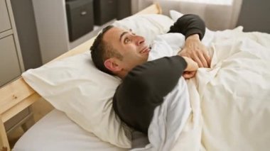 Neşeli bir İspanyol iyi aydınlatılmış bir yatak odasındaki beyaz çarşaflı bir yatakta otururken kutlama yapar..