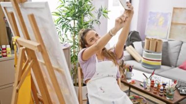 Genç ve güzel İspanyol kadın ressam resim stüdyosunda akıllı telefondan selfie çekiyor.
