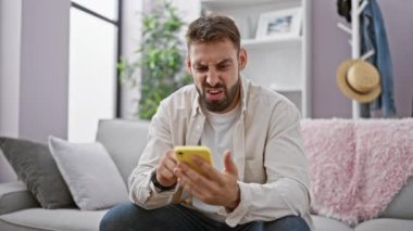 Sakallı, genç, yakışıklı İspanyol adam evdeki kanepede oturmuş, akıllı telefon kullanıyor. Ciddi yetişkin erkek mesajlaşması, kapalı alanda teknolojiye dalmış..