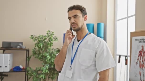 一名身穿白色实验室外套 心事重重的男子在康复诊所的电话中与人交谈 他的解剖海报清晰可见 — 图库视频影像