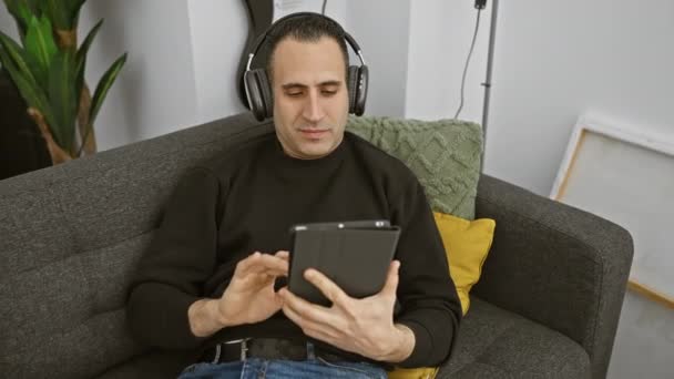 Hispanic Man Avslappnad Klädsel Använder Tablett Soffan Inomhus Med Hörlurar — Stockvideo