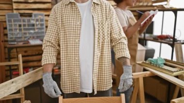 İki profesyonel marangoz, bir erkek ve bir kadın, hareketli işyerlerinde sert duruyor, kolları çapraz bir ortaklık jesti, kereste, marangozluk ve marangozluk aletleri arasında.