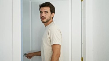 Sakallı genç bir İspanyol, beyaz, modern mobilyalı bir evin içinde dikilirken düşünceli bir şekilde bakıyor..