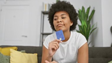 Düşünceli bir kadın elinde kredi kartıyla evde oturur, etrafı ev dekorasyonuyla çevrilidir..