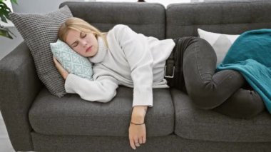 Sarışın kadın rahat bir evin içinde yastık ve turkuaz battaniyeyle koltukta dinleniyor..