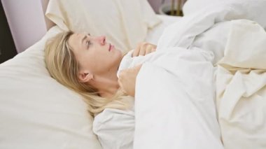 Genç sarışın bir kadın yatak odasında endişeli görünüyor, beyaz çarşaflarını içeride tutuyor..