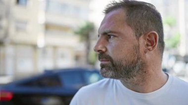 Gri saçlı, sakallı, güneşli bir sokak ortamında gülümseyen orta yaşlı yakışıklı bir adam..